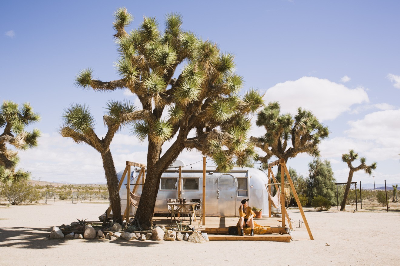 Coachella outfit ideas by Los Angeles Fashion Blogger Laura Lily | How to Take Good Travel Photos by popular California travel blogger, Laura Lily: image of a woman sitting under a Joshua Tree with an airstream trailer in the distance.
