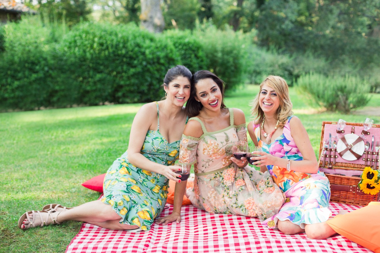 Summer Picnic in Italy by Lifestyle Blogger Laura Lily, I Am Fashion Laine, Elizabeth Keene | An Italian Picnic by popular US fashion blogger, Laura Lilly: image of 3 women in garden dresses sitting together on a red and white checked picnic blanket and holding glasses of red wine at La Foce Property in Tuscany, Italy.