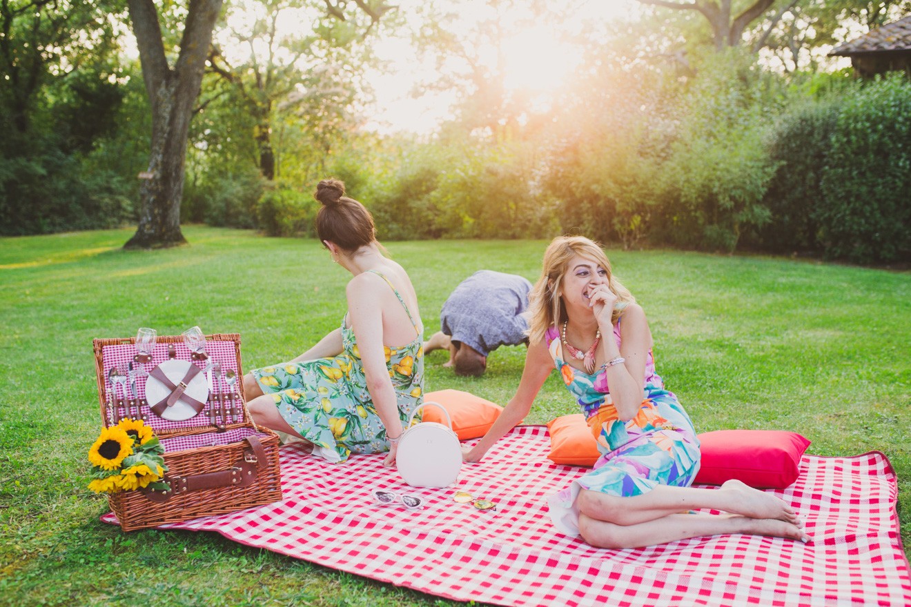 Summer Picnic in Italy by Lifestyle Blogger Laura Lily, I Am Fashion Laine, Elizabeth Keene | An Italian Picnic by popular US fashion blogger, Laura Lilly: image of 2 women in garden dresses sitting together on a red and white checked picnic blanket, laughing and watching a little boy do a summersault at La Foce Property in Tuscany, Italy