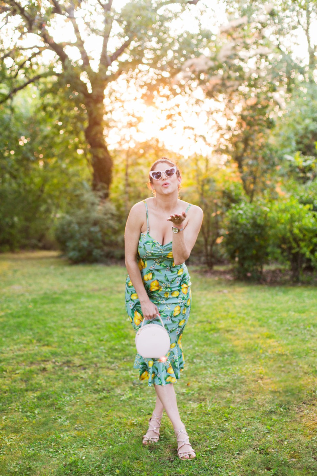 Summer Picnic in Italy by Lifestyle Blogger Laura Lily, I Am Fashion Laine, Elizabeth Keene | An Italian Picnic by popular US fashion blogger, Laura Lilly: image of a woman in a lemon print boohoo dress at La Foce Property in Tuscany, Italy wearing heart shaped sunglasses and blowing a kiss.
