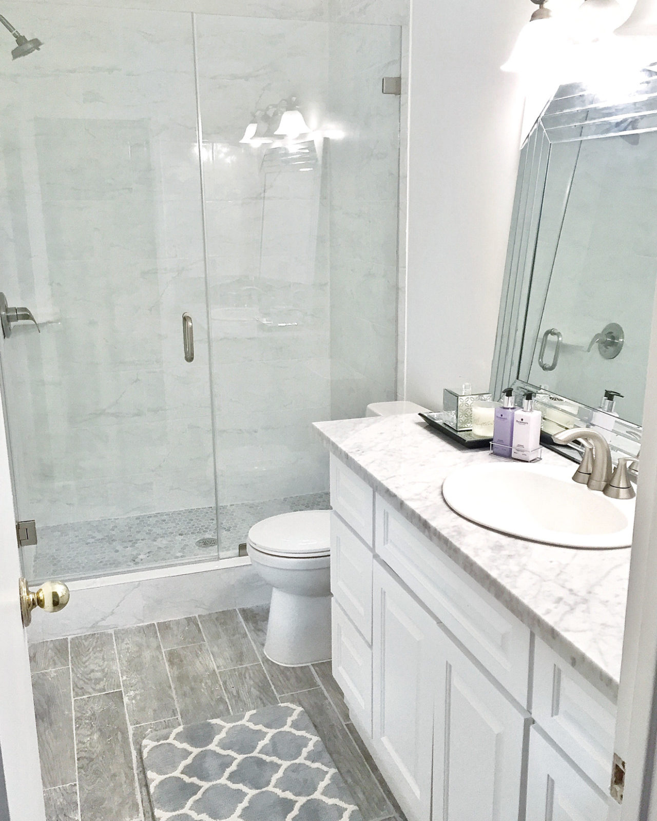 Modern Bathroom Remodel Ideas + Reveal featured by popular US lifestyle blogger, Laura Lily: image of after photo of modern bathroom remodel with gray ceramic faux wood tiles, glass shower doors, gray hexagon tile, marble counter, marble subway tile, custom vanity, and purple towels and rug.