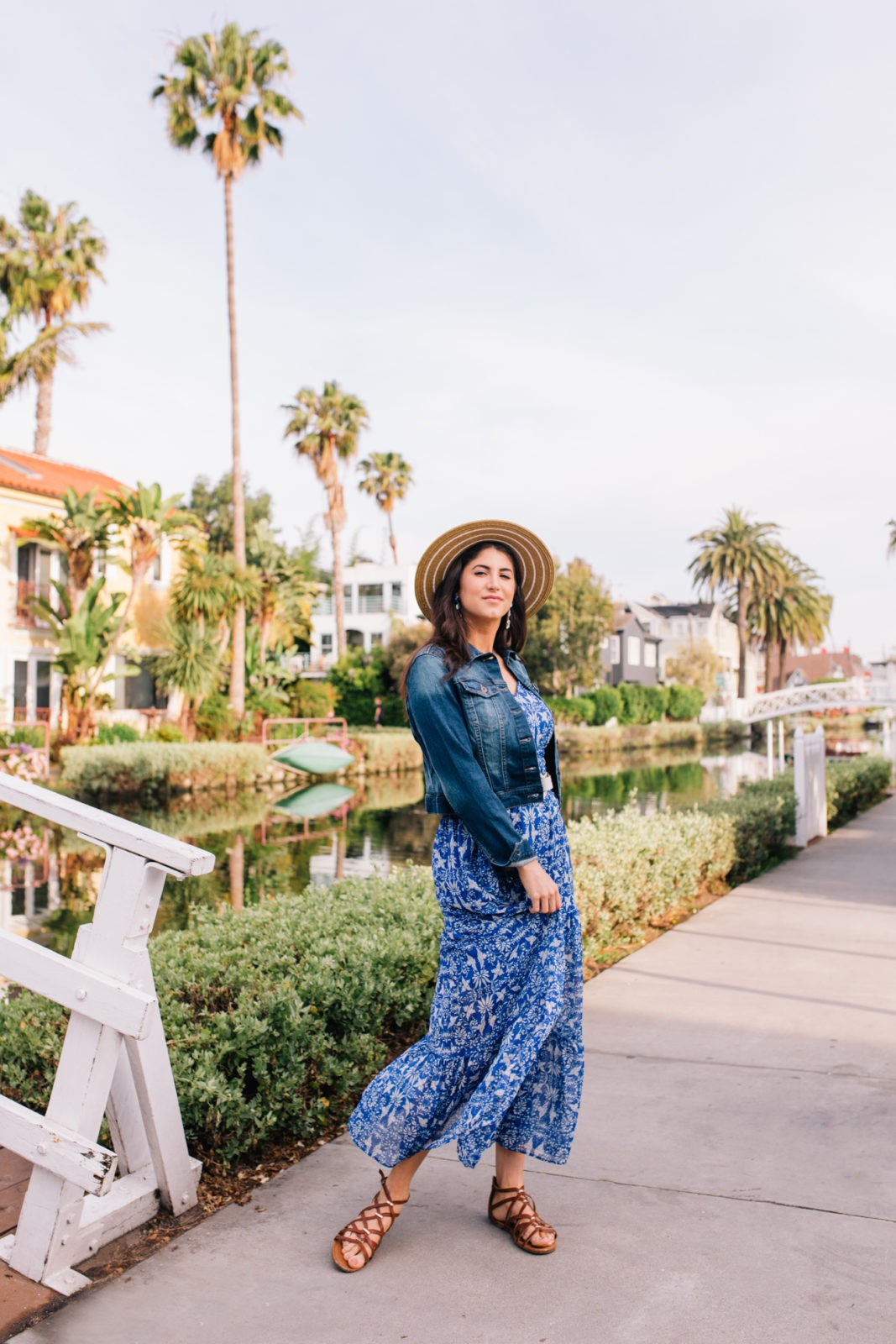 Stage Summer Moments, 2019 Summer Bucket List Moments by popular Los Angeles Lifestyle Blogger Laura Lily: image of woman at Venice, California canals wearing Stage Stores flowing blue and white floral maxi dress, gladiator sandals, cropped jean jacket and straw sun hat. 