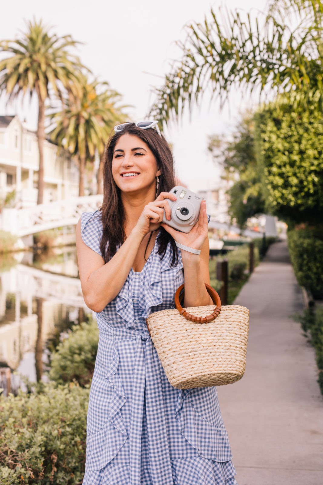 Stage Summer Moments, 2019 Summer Bucket List Moments by popular Los Angeles Lifestyle Blogger Laura Lily: image of woman next to Venice, California canals holding a gray Polaroid Instax camera and wearing a blue and white gingham ruffle wrap dress, white sunglasses and holding a straw tote. 