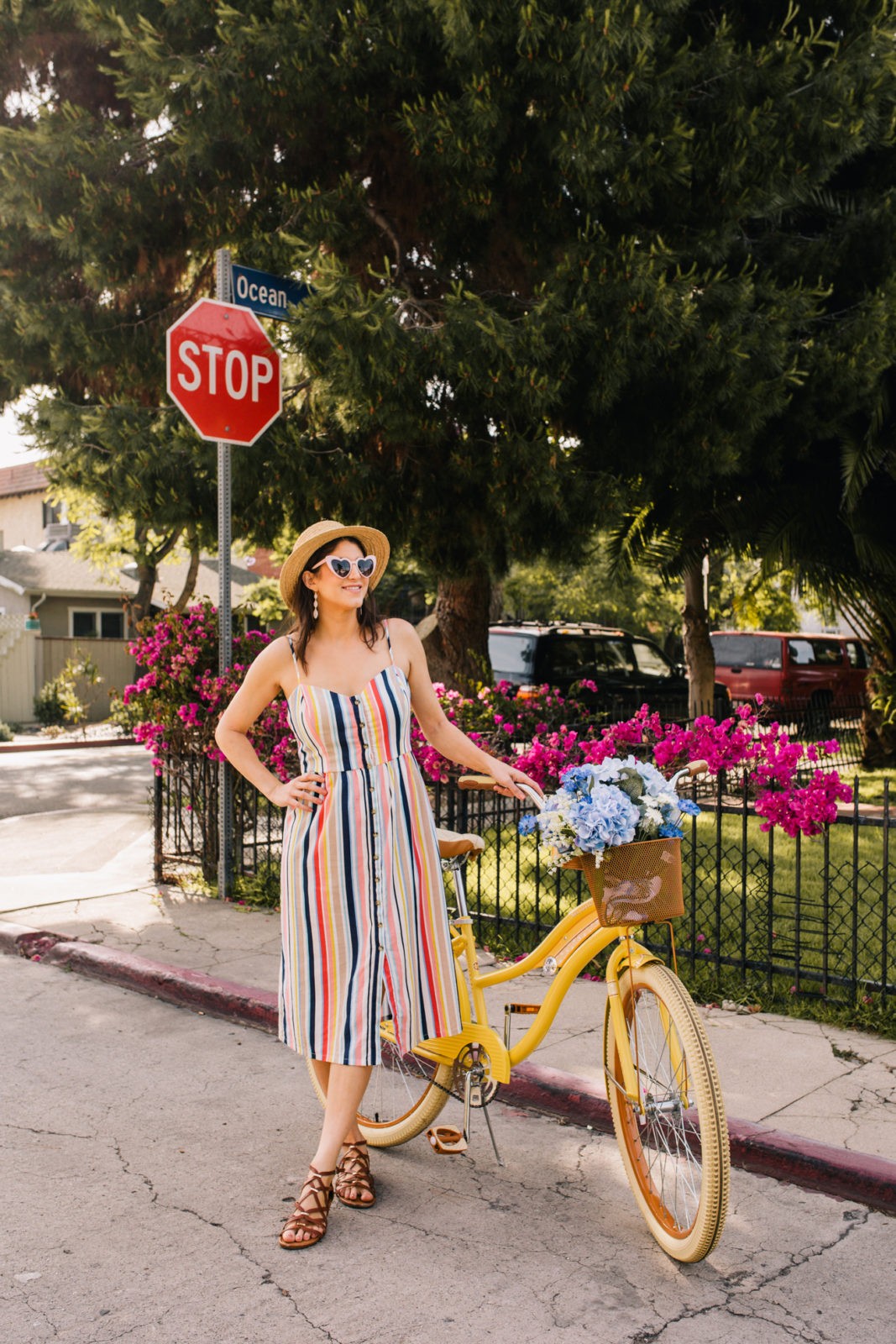 Stage Summer Moments, 2019 Summer Bucket List Moments by popular Los Angeles Lifestyle Blogger Laura Lily: image of woman standing next to Stage Stores yellow beach cruiser bike with bike basket full of flowers while wearing multi colored stripe spaghetti stripe dress, straw sun hat, brown gladiator sandals and pink heart shaped sunglasses.