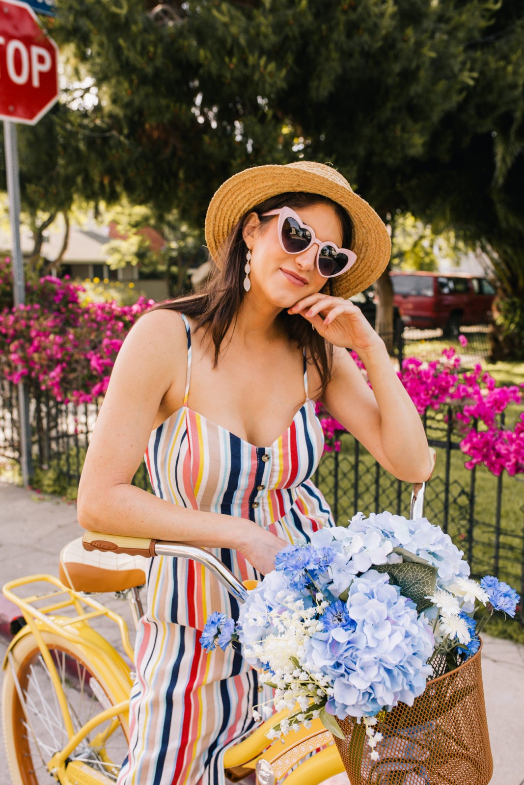 Stage Summer Moments, 2019 Summer Bucket List Moments by popular Los Angeles Lifestyle Blogger Laura Lily: image of woman on Stage Stores yellow beach cruiser bike with bike basket full of flowers while wearing multi colored stripe spaghetti stripe dress, straw sun hat, and pink heart shaped sunglasses.