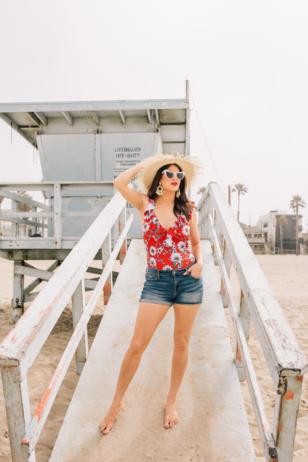 Stage Summer Moments, 2019 Summer Bucket List Moments by popular Los Angeles Lifestyle Blogger Laura Lily: image of woman standing on lifeguard tower ramp at the beach and wearing a Stage Stores red daisy print one piece swimsuit with ruffle details, white heart shaped sunglasses, straw sun hat, tassel earrings, and cutoff denim shorts. 