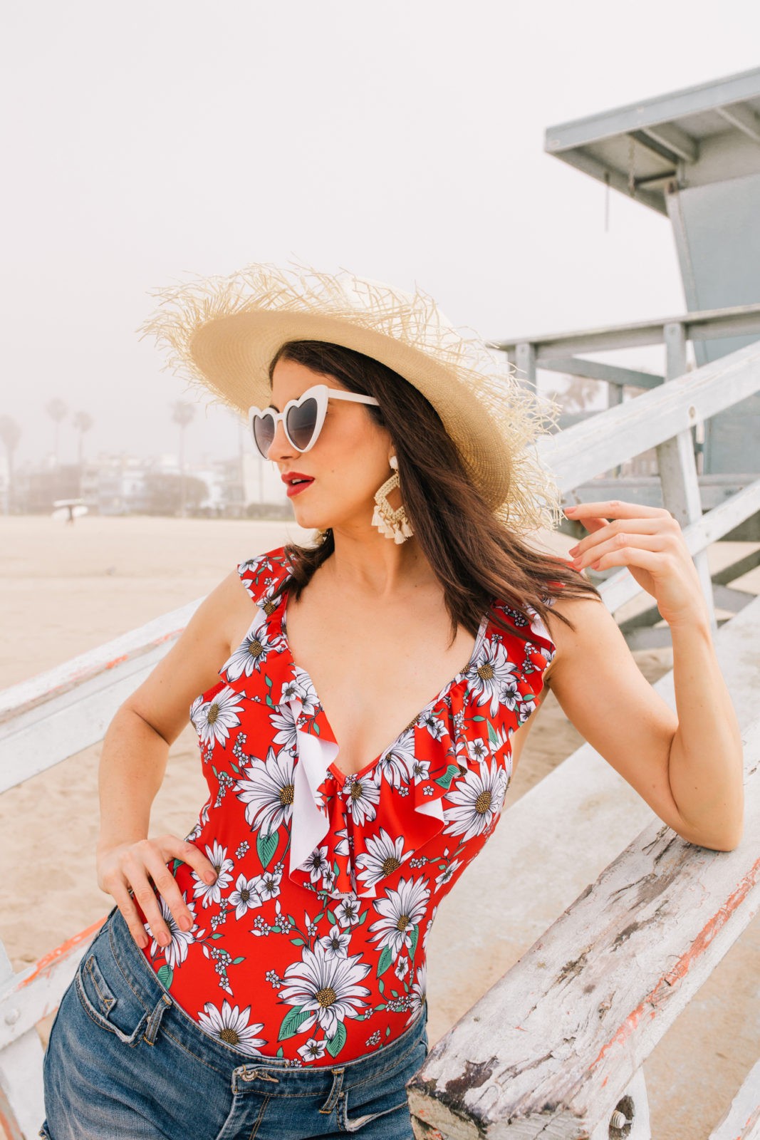 Stage Summer Moments, 2019 Summer Bucket List Moments by popular Los Angeles Lifestyle Blogger Laura Lily: image of woman standing on lifeguard tower ramp at the beach and wearing a Stage Stores red daisy print one piece swimsuit with ruffle details, white heart shaped sunglasses, straw sun hat, tassel earrings, and cutoff denim shorts. 