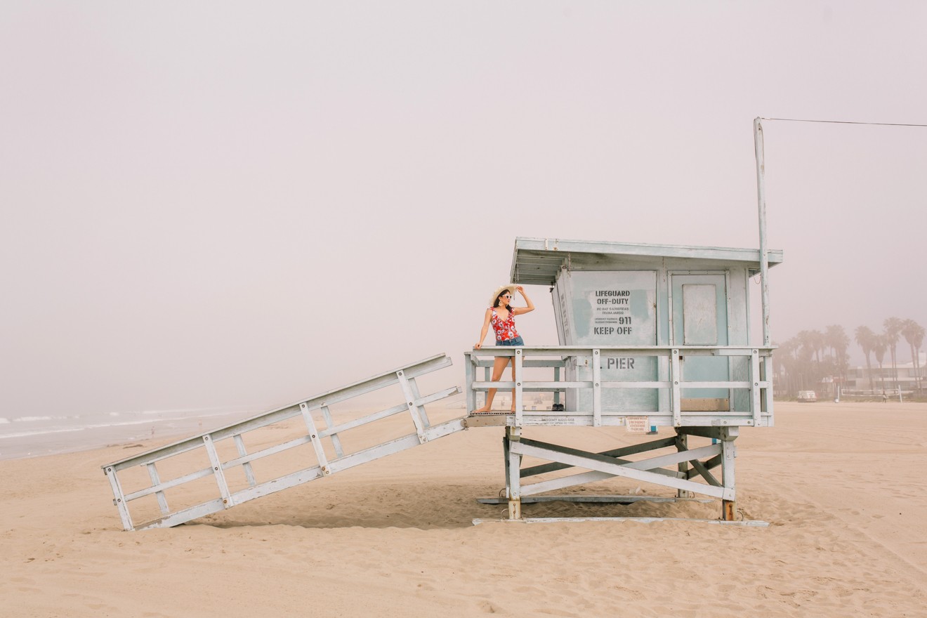 Stage Summer Moments, 2019 Summer Bucket List Moments by popular Los Angeles Lifestyle Blogger Laura Lily: image of woman standing on lifeguard tower at the beach and wearing a Stage Stores red daisy print one piece swimsuit with ruffle details, white heart shaped sunglasses, straw sun hat, tassel earrings, and cutoff denim shorts. 