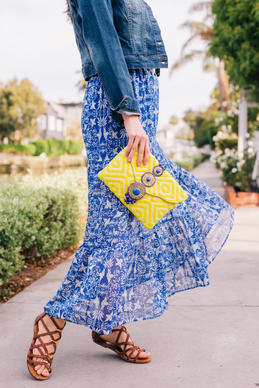 Stage Summer Moments, 2019 Summer Bucket List Moments by popular Los Angeles Lifestyle Blogger Laura Lily: image of woman at Venice, California canals wearing Stage Stores flowing blue and white floral maxi dress, brown gladiator sandals, cropped jean jacket and holding a bright yellow clutch 