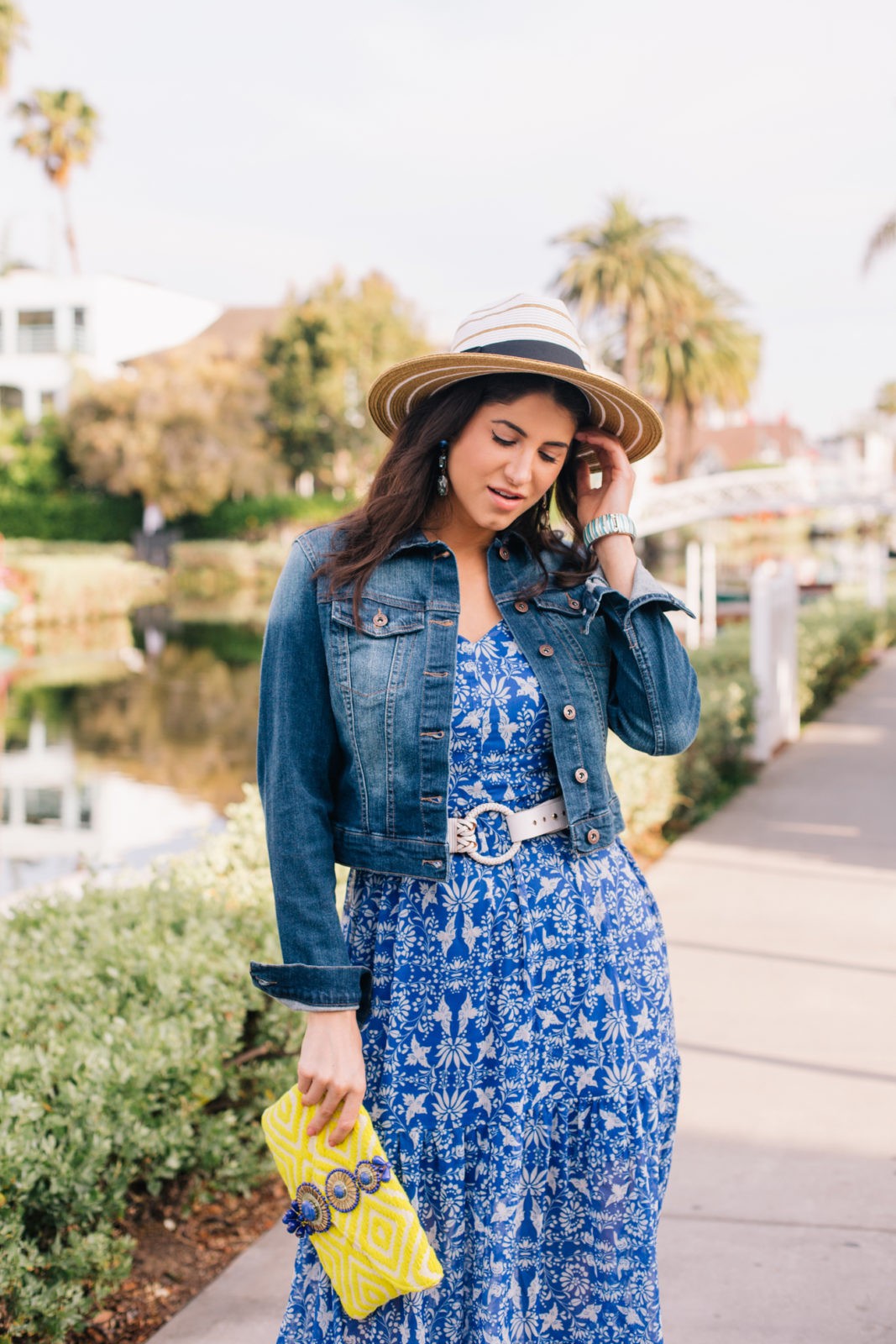 Stage Summer Moments, 2019 Summer Bucket List Moments by popular Los Angeles Lifestyle Blogger Laura Lily: image of woman at Venice, California canals wearing Stage Stores flowing blue and white floral maxi dress, white belt, cropped jean jacket, straw sun hat and holding a yellow clutch. 
