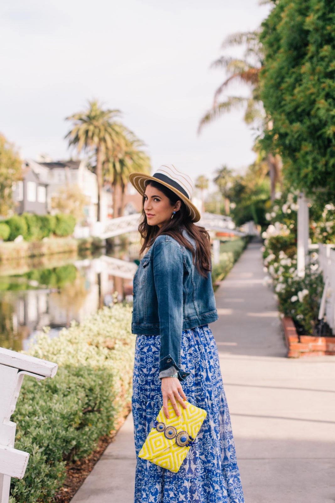 Stage Summer Moments, 2019 Summer Bucket List Moments by popular Los Angeles Lifestyle Blogger Laura Lily: image of woman at Venice, California canals wearing Stage Stores flowing blue and white floral maxi dress, cropped jean jacket, straw sun hat and holding a yellow clutch.