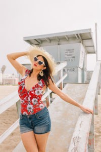 Cute Summer Fringe Hats featured by top US fashion blogger, Laura Lily: image of a woman at the beach wearing a Tenth Street fringe hat, heart shaped sunglasses, Jessica Simpson floral one piece swimsuit, Jessica Simpson denim cuff shorts.