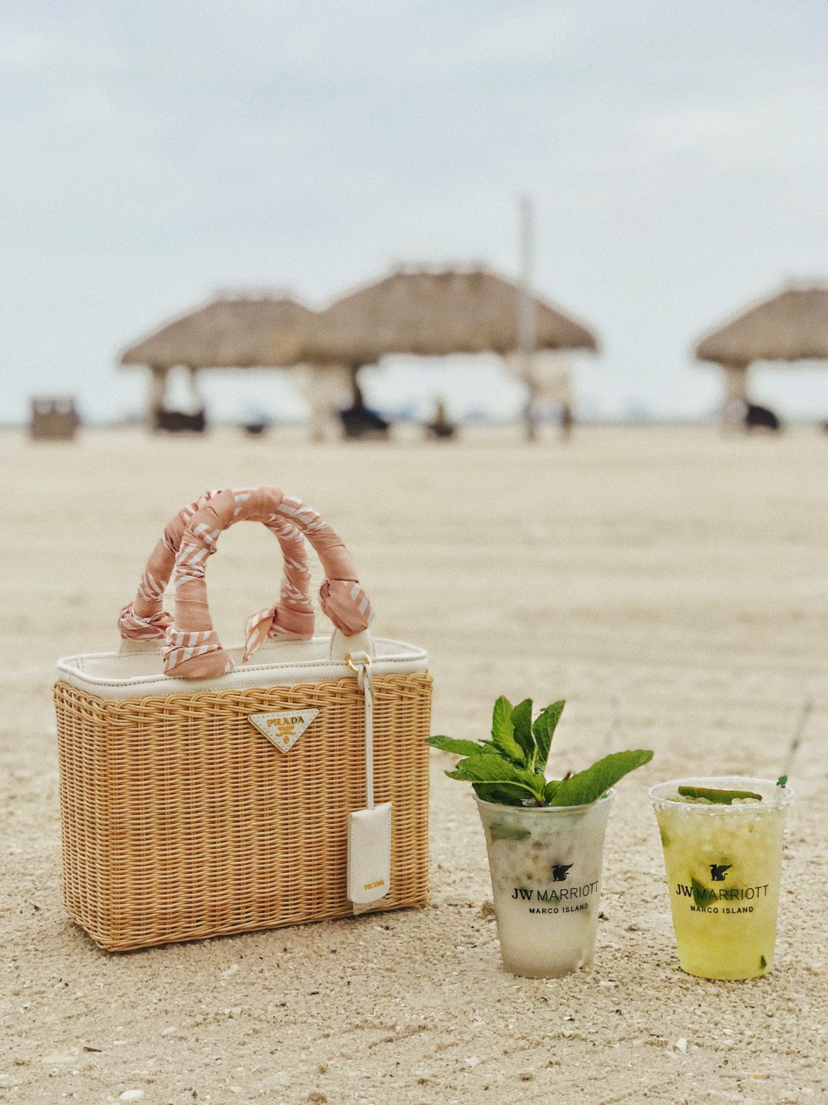 JW Marriott Marco Island by Luxury Travel Blogger Laura Lily: image of prada white & beige raffia garden tote resting in the sand on the beach next to a cocktail.