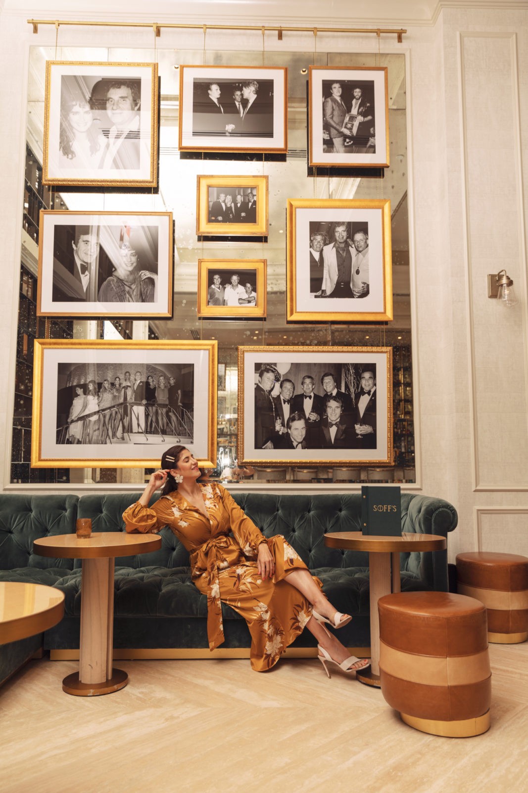 JW Marriott Turnberry Resort Miami by popular travel blogger, Laura Lily: image of a woman sitting in the Miami Turnberry Resort on a velvet tuft couch under a wall full of black and white photos in gold picture frames.