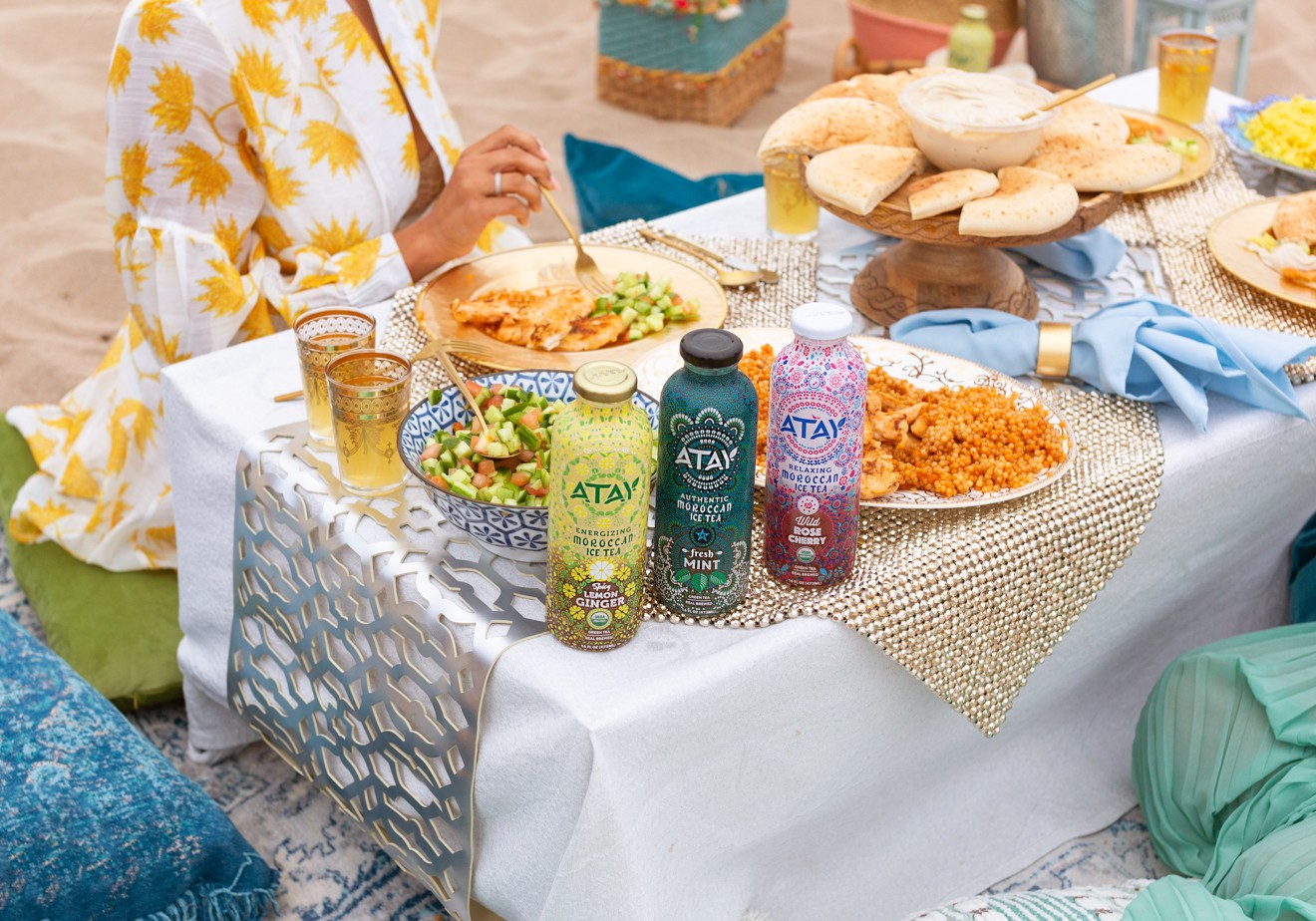 Moroccan Picnic with Atay Tea by popular California life and style blogger, Laura Lily: image of woman sitting in the sand for a moroccan picnic at the beach with gold beaded placemats, various moroccan foods, blue pillows, blue linen napkins, gold embellished moroccan cups, gold plate, gold silverware, and Atay Tea.