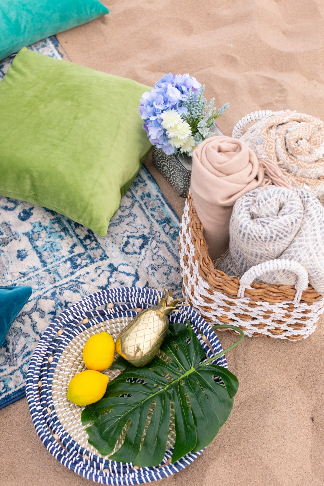 Moroccan Picnic with Atay Tea by popular California life and style blogger, Laura Lily: image of a woven basket with a palm front, lemons, and gold pineapple drink mixer, a woven basket filled with throw blankets, a blue oriental rug, and blue and green throw pillows.
