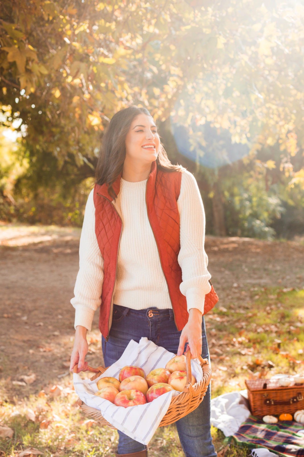 Cozy Fall Favorites by Los Angeles Fashion Blogger Laura Lily | Cozy Fall Fashion Trends by popular California fashion blogger, Laura Lily: image of a woman holding a basket of apples outside and wearing a H&M Chenille Sweater, jeans, Born Boots, and a quilted vest.